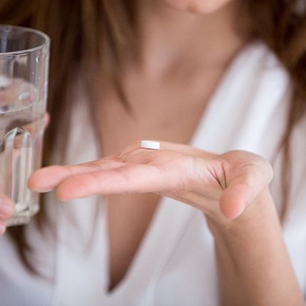 Woman in white holding single small pill in the palm of one hand and glass of water with other hand