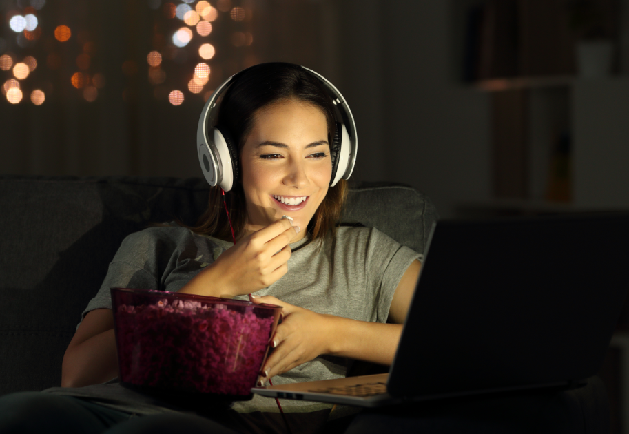 Woman eating popcorn and watching laptop with headphones on, smiling